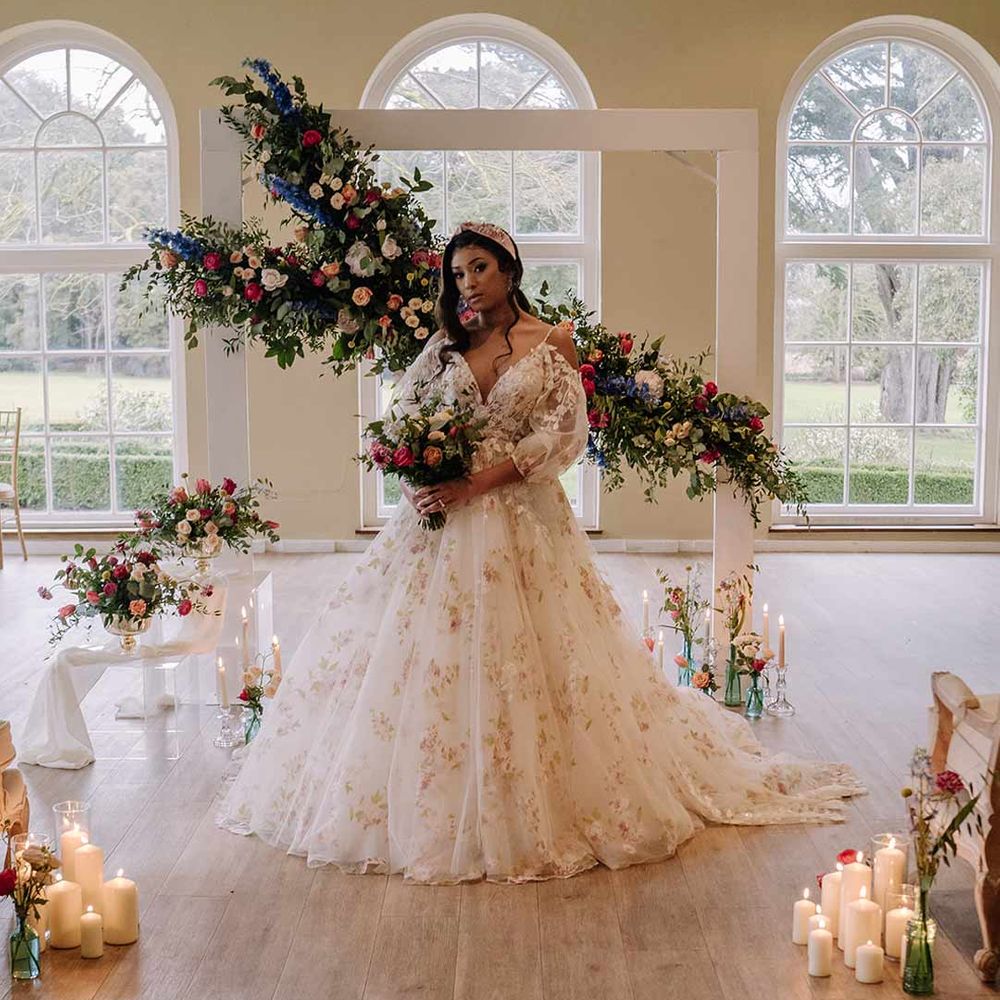 Bride in v-neck long sleeve shoulder cut out 3d appliqué wedding dress with flecks of gold wearing satin bridal headband holding large dried flower, eucalyptus, spring florals and garden rose bridal bouquet by suspended spring floral decorations and pillar candles at Braxted Park wedding venue 