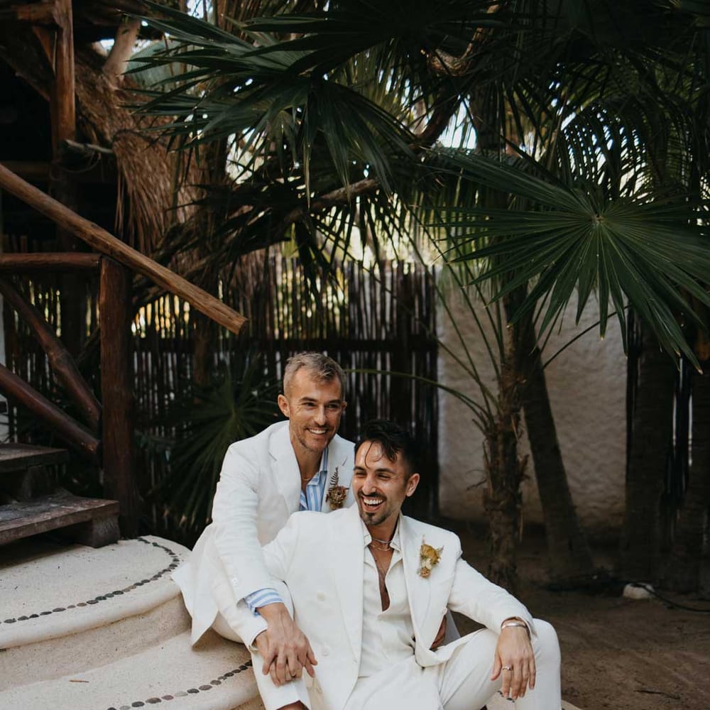 Groom sitting on stairs of hotel wedding venue wearing white blazer with dusky orange garden rose and dried flower boutonniere, white shirt, white suit trousers and silver jewellery with groom in white mens wedding suit,  light blue and white striped shirt, dusky orange garden rose and dried flower boutonniere, white suit trousers and gold jewellery 