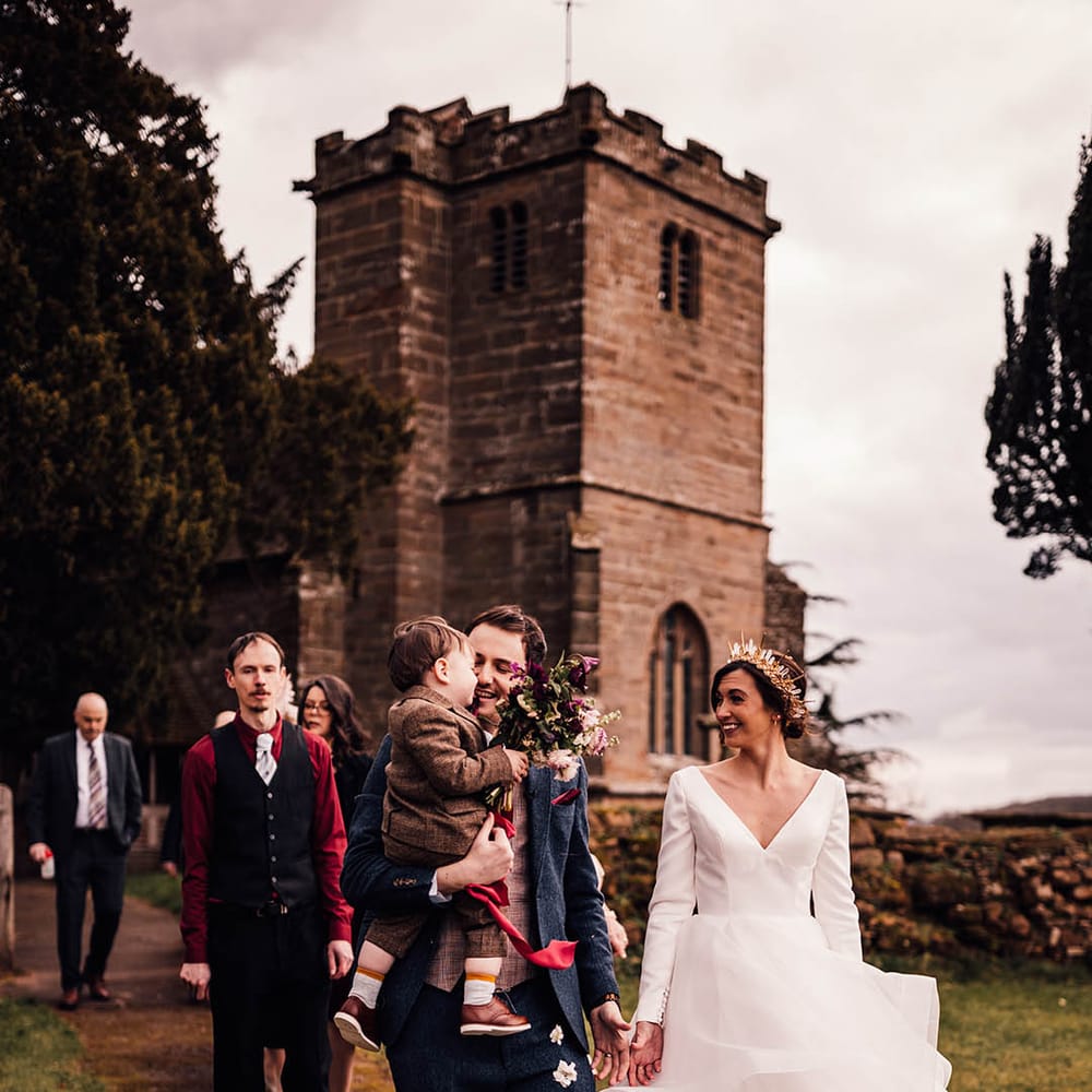 Groom in navy three piece suit with red tie carrying their toddler in a brown suit walking with the bride in a traditional princess style wedding dress