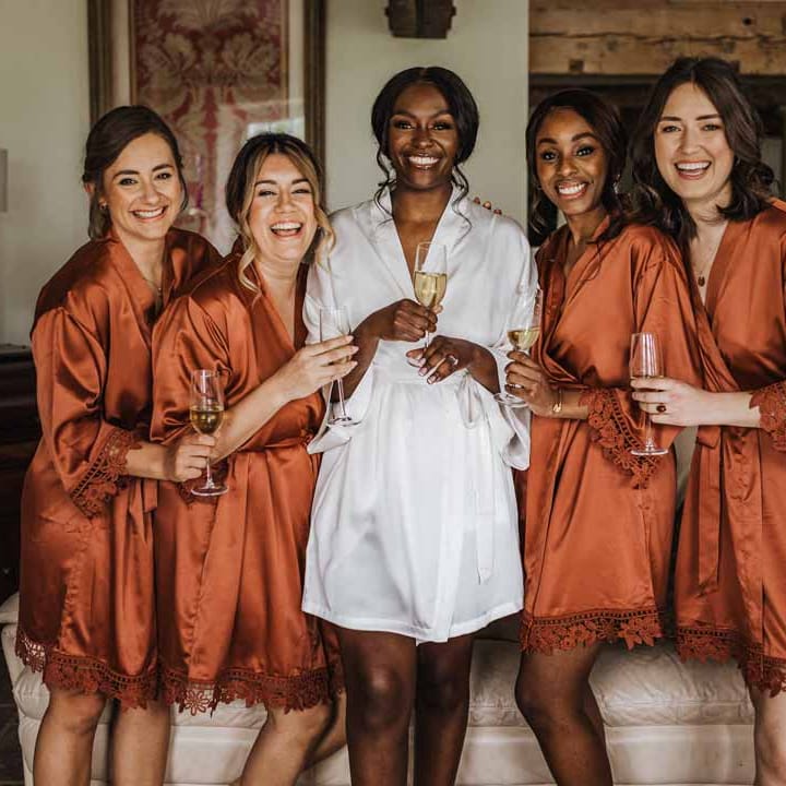 Bride in white satin getting ready robe with bridal party wearing burnt orange getting ready robes at Lains Barn wedding venue 