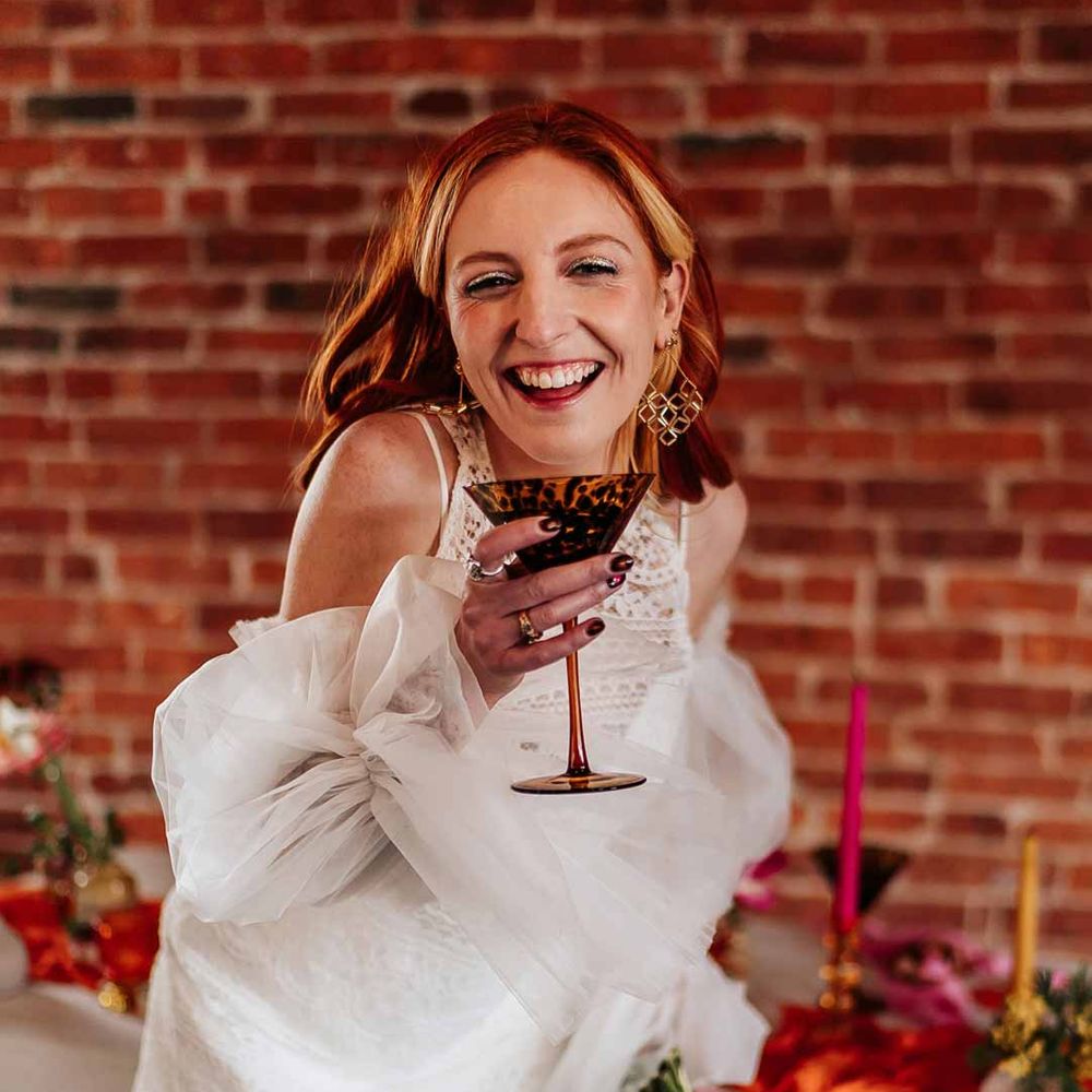 Bride in 70s inspired boatneck short lace wedding dress with detachable tulle puff sleeves and platform gold wedding heels sipping from leopard print cocktail glass holding colourful mixed flower bridal bouquet 