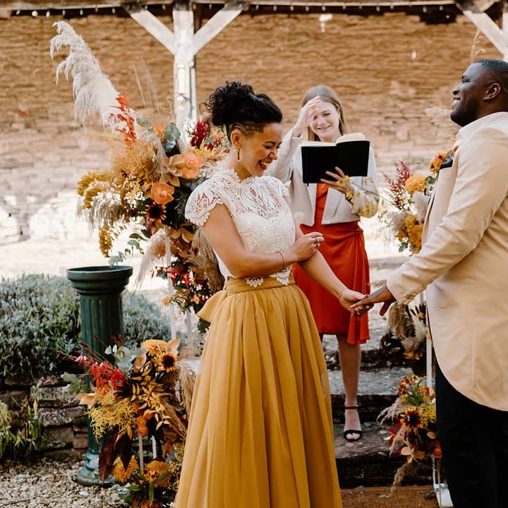 Groom in cream morning suit with forest green grooms waistcoat and trousers, forest green pocket square and orange boutonniere standing and holding hands with bride in white mesh lace bridal crop top and yellow wedding skirt