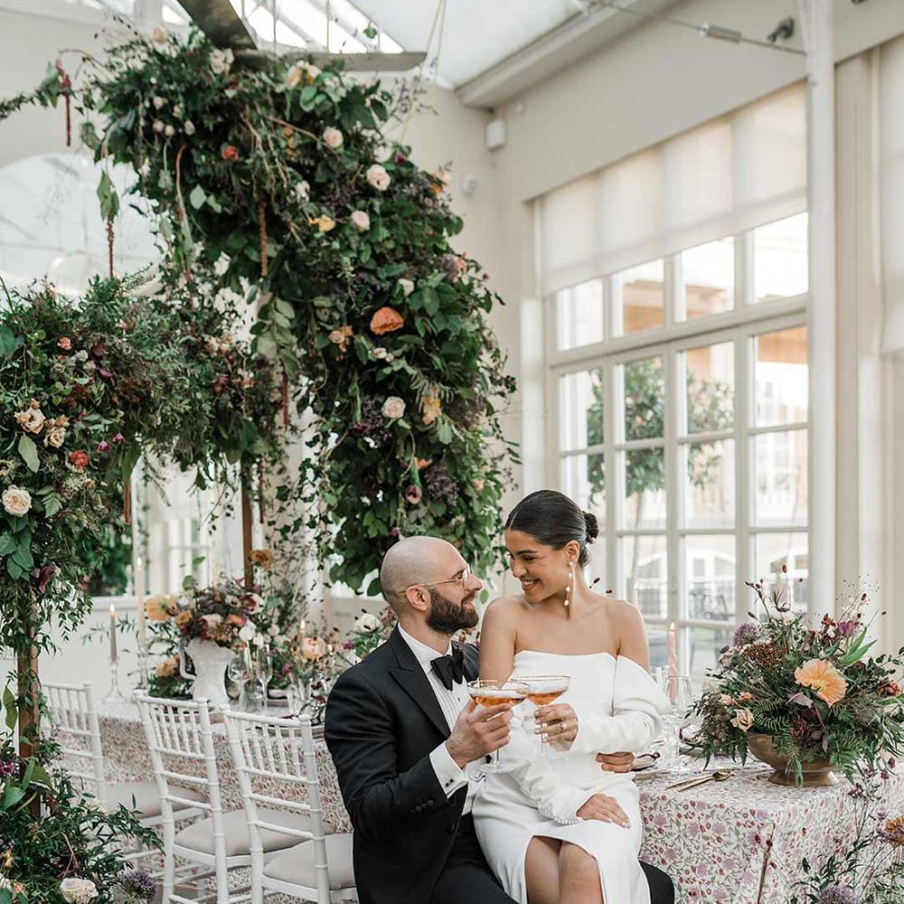Groom in classic black tuxedo with bowtie and wildflower boutonniere with bride on his lap wearing long sleeve off the shoulder wedding dress with pearl button detailing and a side slit holding cocktail sitting by botanical wedding tablescape with patterned floral tablecloth, twisted tapered candles, art deco vases with dried wildflower wedding centrepieces surrounded by suspended wildflower and foliage arrangements in large gold vases at Buxted Park