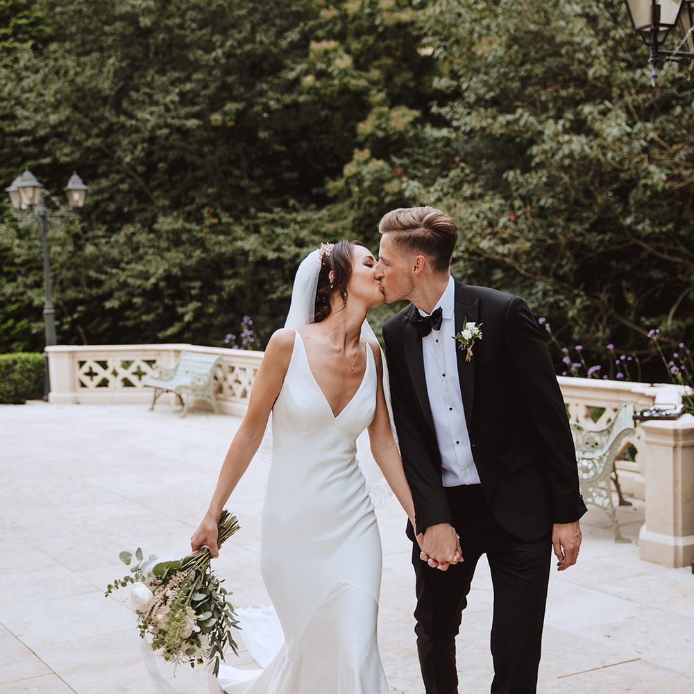 Bride in plunging satin slip wedding dress with cathedral length veil kissing groom in black tie 
