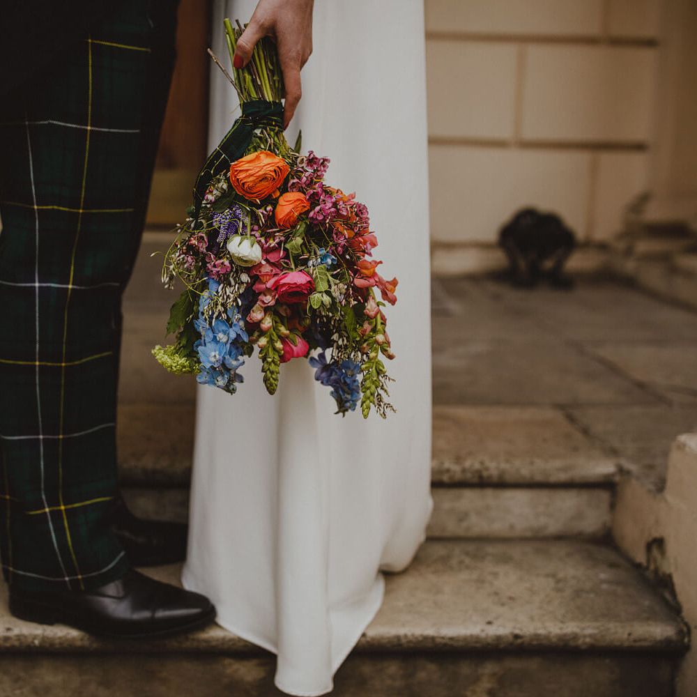 Bride and groom facing each other, bride holding bright poppy bouquet against white bridal gown, groom wears tartan trousers