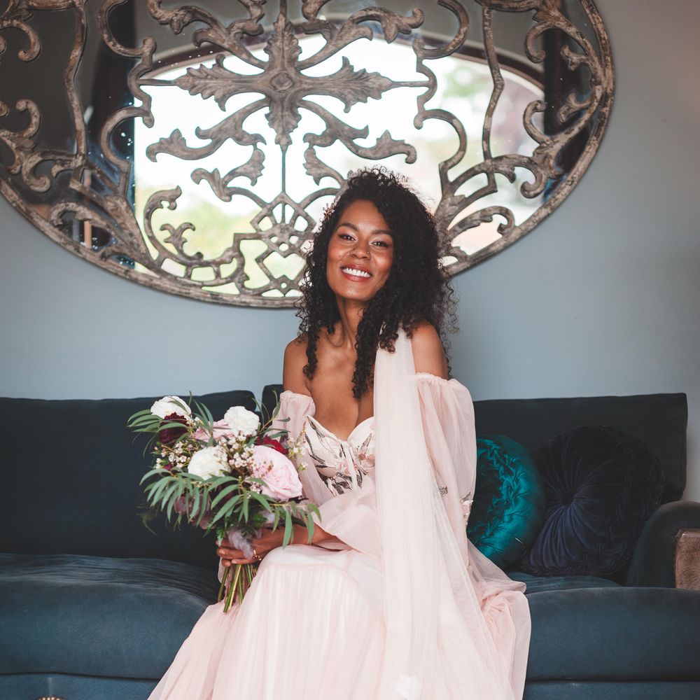 Black bride with naturally curly hair in blush bridal separates with an applique veil sitting on a sofa holding a deep red. pink, white and green bouquet