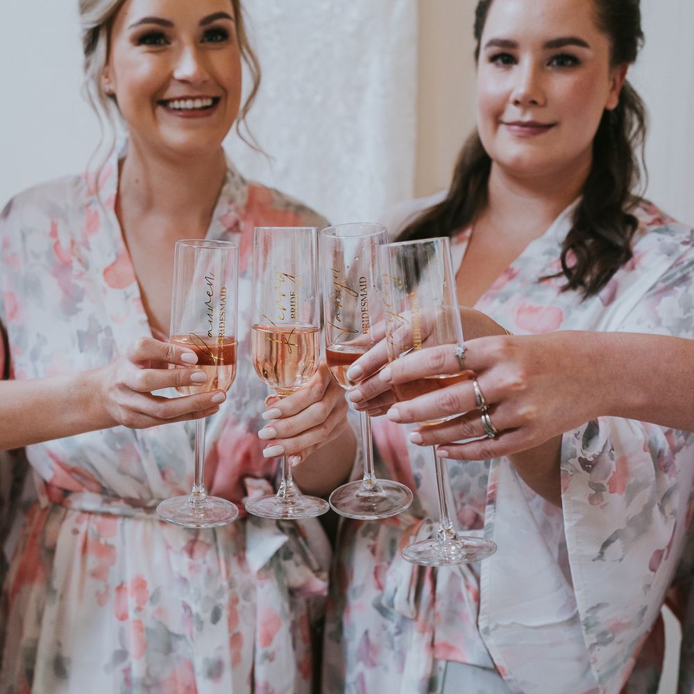 Bride and bridesmaids in floral bridal gowns clinking personalised flutes before Tythe Barn wedding with barn wedding flowers