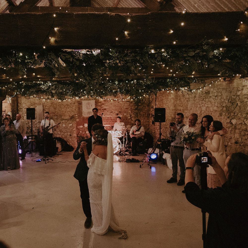 Bride in Grace Loves Lace bardot wedding dress and veil dances with groom in blue suit jacket under fairy light and foliage canopy as guests watch on at Isle of Wight wedding with macrame wedding decor