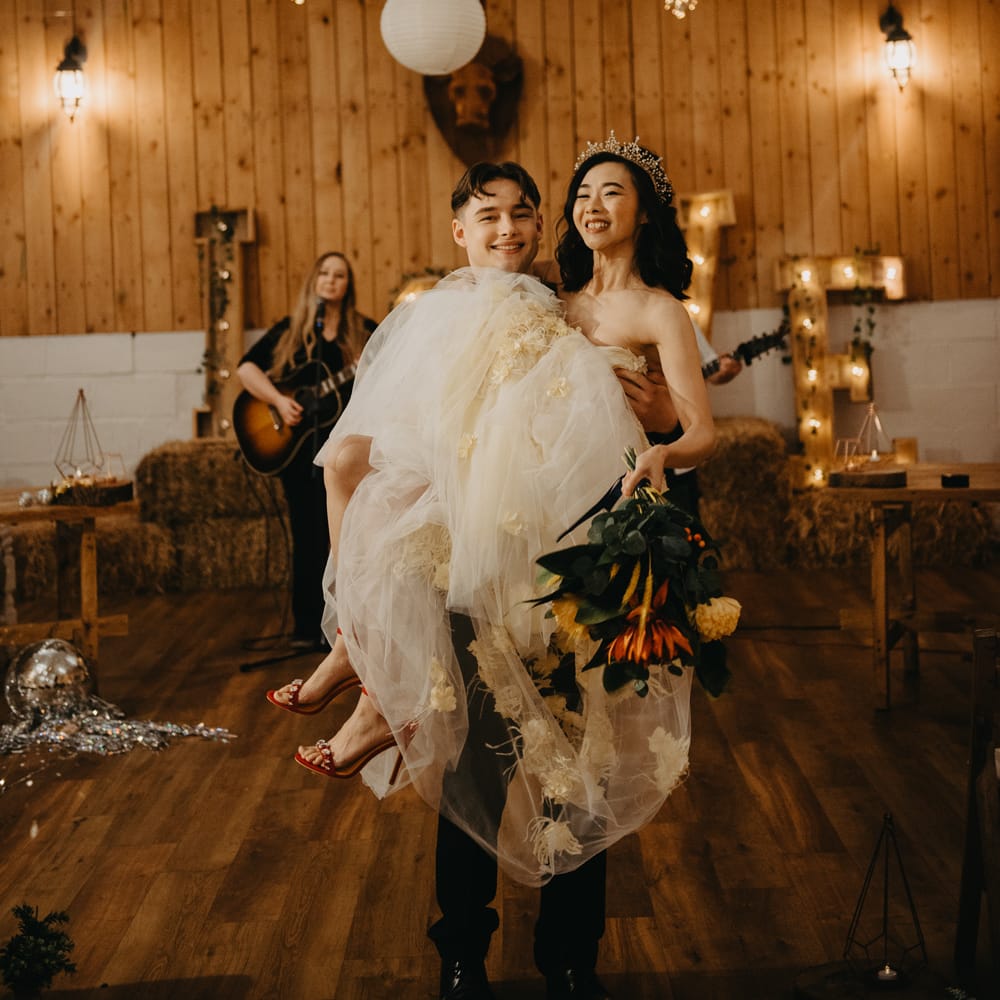 Bride in feathered tulle wedding dress and red heels holding bouquet is held by groom in dark suit during rustic wedding ceremony with fairy lights