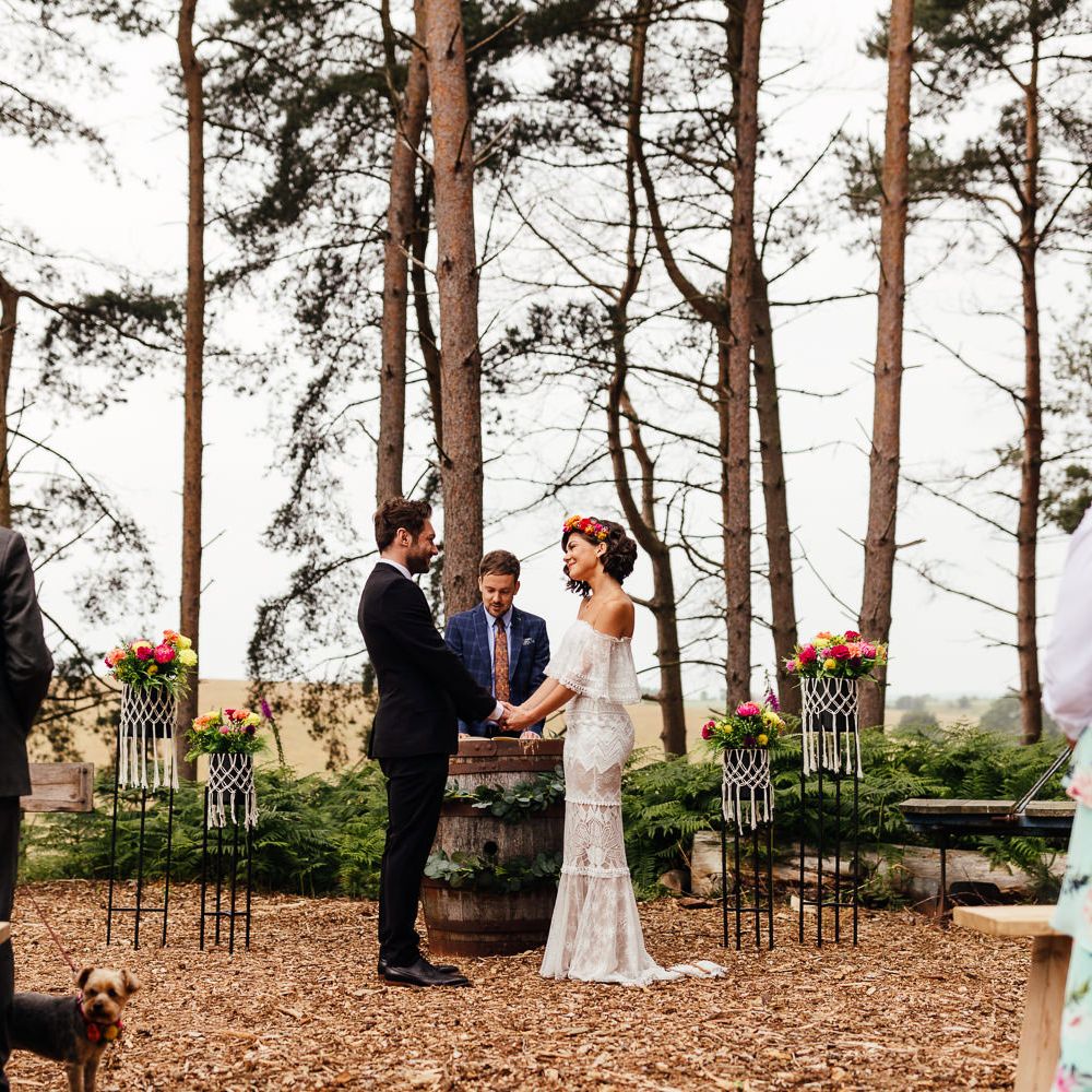 Bride in Grace Loves Lace wedding dress and flower crown stands holding hands with groom in black suit during woodland wedding ceremony at The Bridal Barn wedding venue