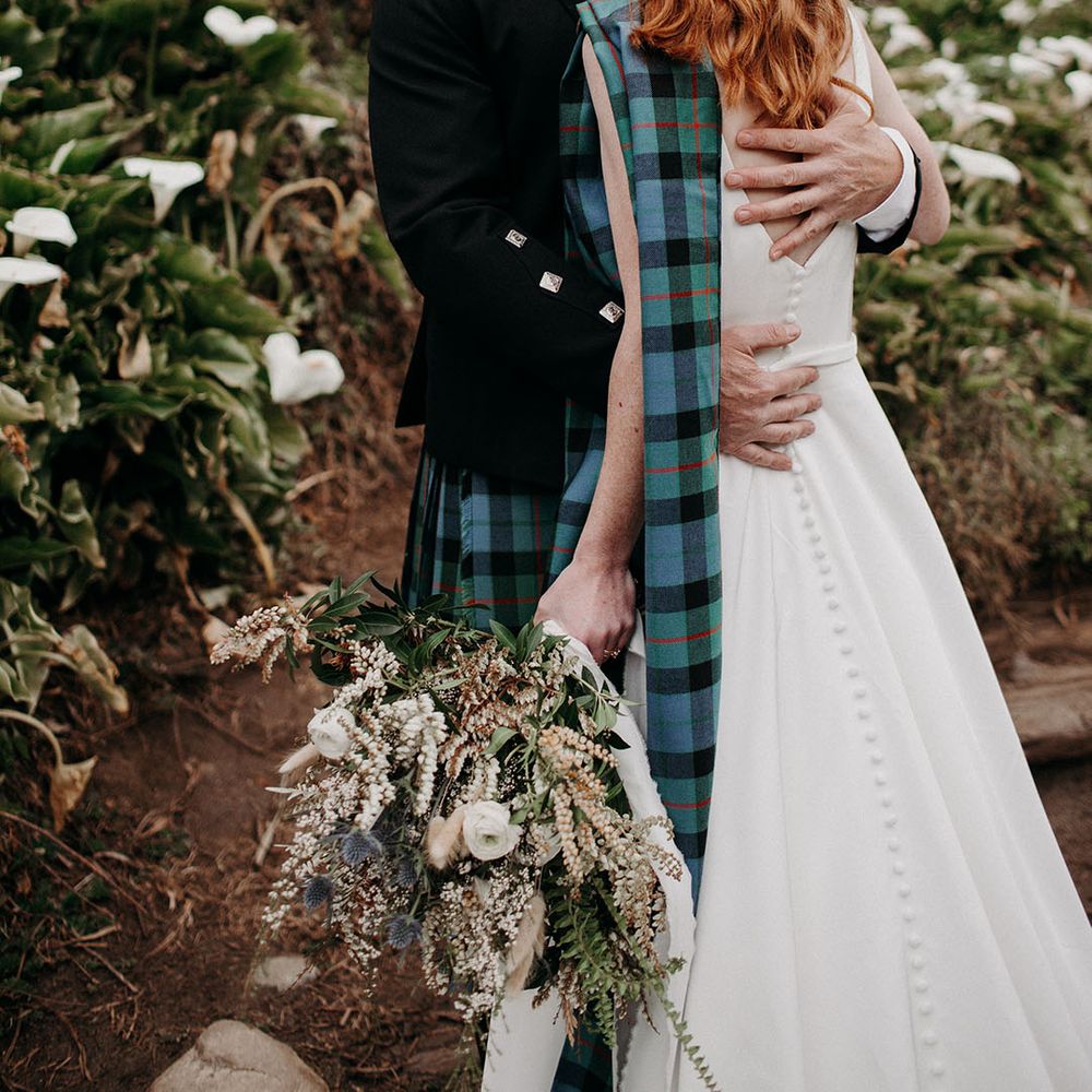 Groom embracing his bride with half up half down red hair holding an anemone and snowdrop wedding bouquet 