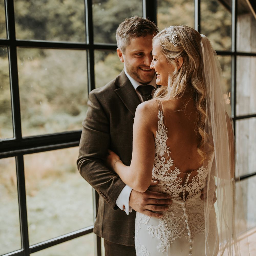 Bride in low back lace wedding dress with button detailing and veil stands with groom in brown suit by large window for rustic wedding in the Lake District