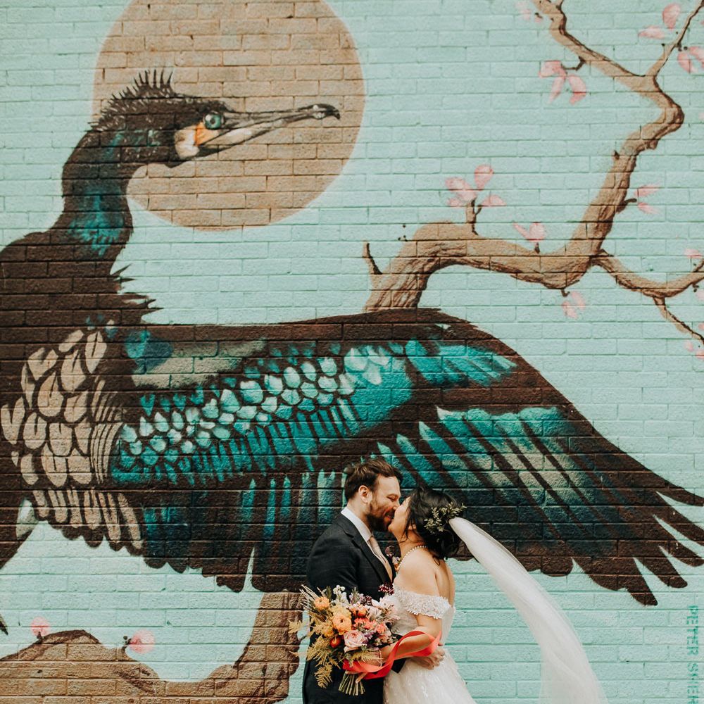 Bride in lace wedding dress with train and veil holding colourful bouquet kisses groom in check suit and brown shoes in front of large street art painting of a bird in Liverpool 