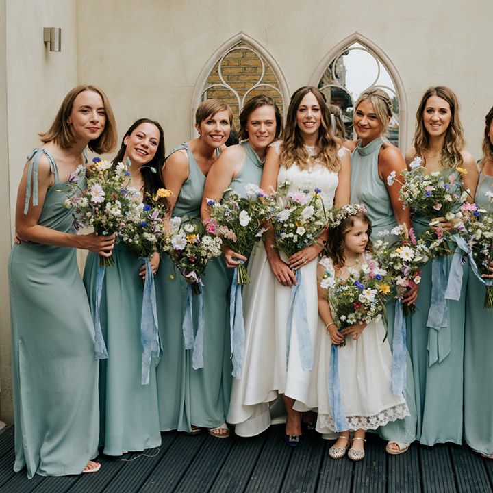 Bride with all her bridesmaids in sage green dresses with varied necklines and wild flower bouquets tied with pale blue ribbon