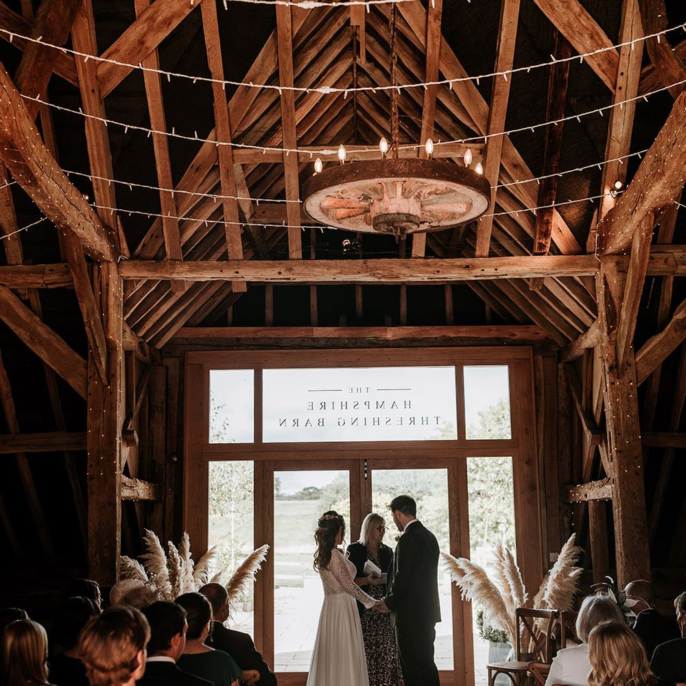 Bride and groom stand at the altar of their rustic barn wedding venue with fairy lights, pampas grass and wooden seats