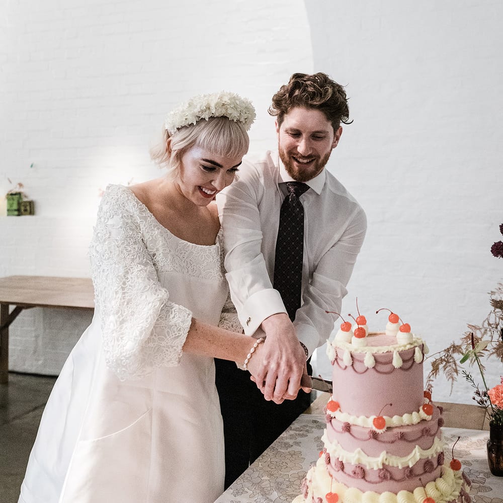 Bride in short wedding dress and groom in patterned tie cut their three tier purple and white wedding cake decorated with cherries and cupcakes
