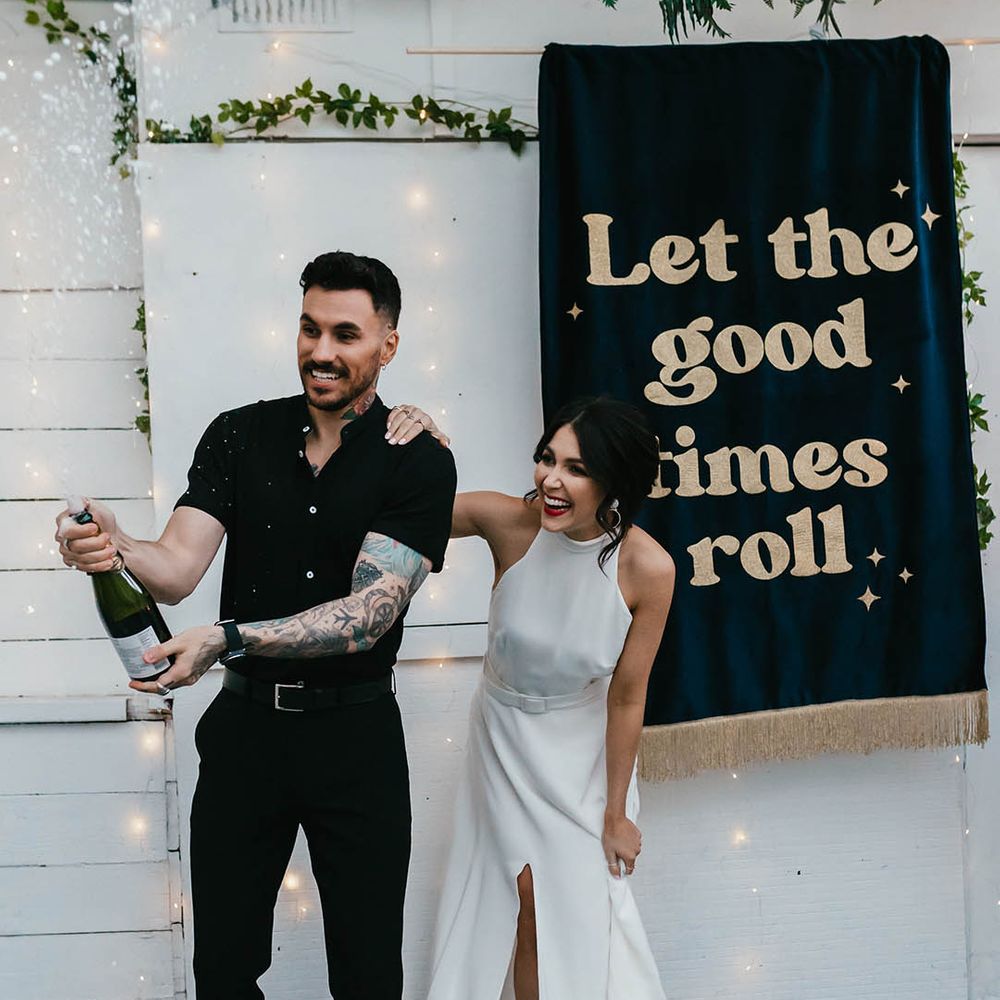 Bride in halter neck wedding dress and groom in black shirt doing champagne pop