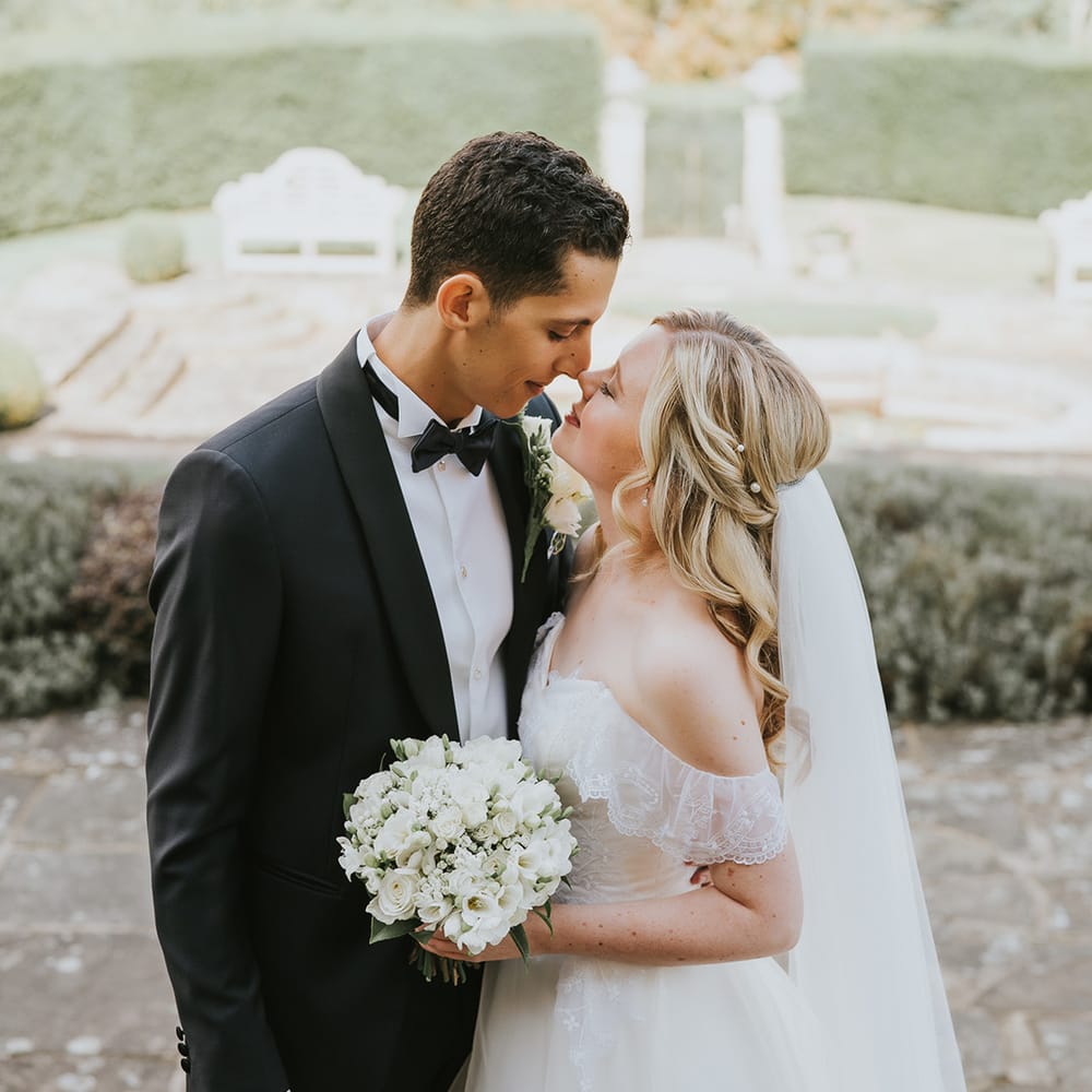 Cute bride and groom couple portrait with bride in lace off the shoulder wedding dress with pearl hair pins and white bouquet with groom in black tie 
