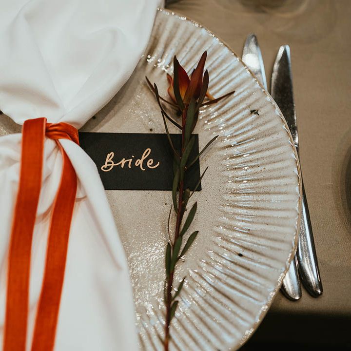 White and silver wedding plate with white napkin tied with burnt orange ribbon with black name place card and flower