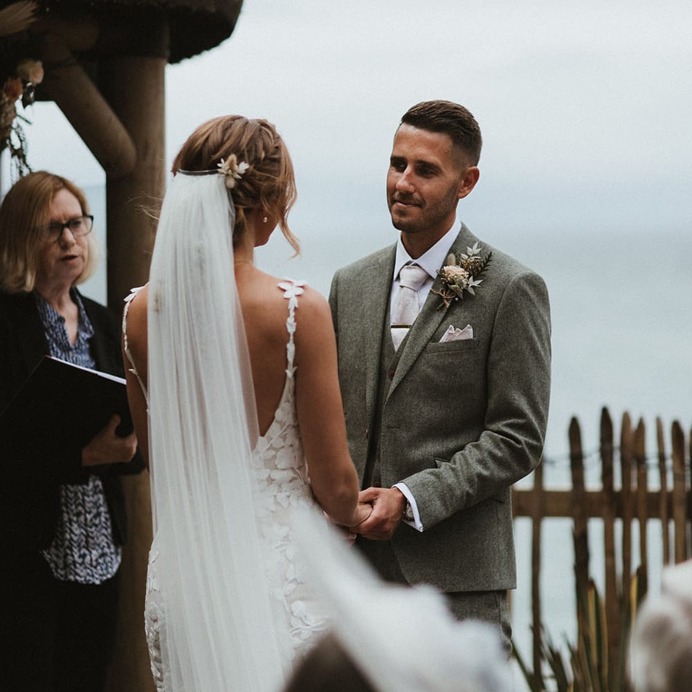 Bride wears floor length veil pinned into her fishtail plaited hair as she and her groom say their vows during outdoor wedding ceremony