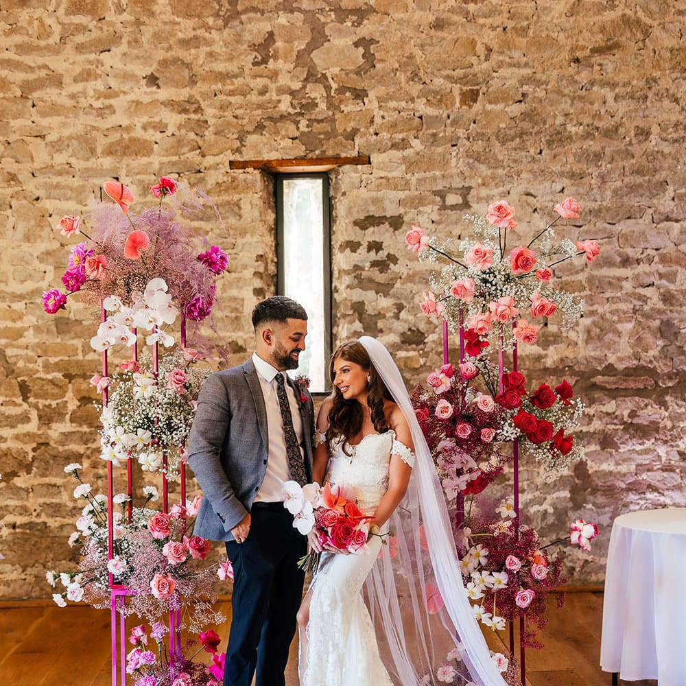 Bride wears lace wedding dress whilst stood with her groom in grey suit in front of a bright floral arch 