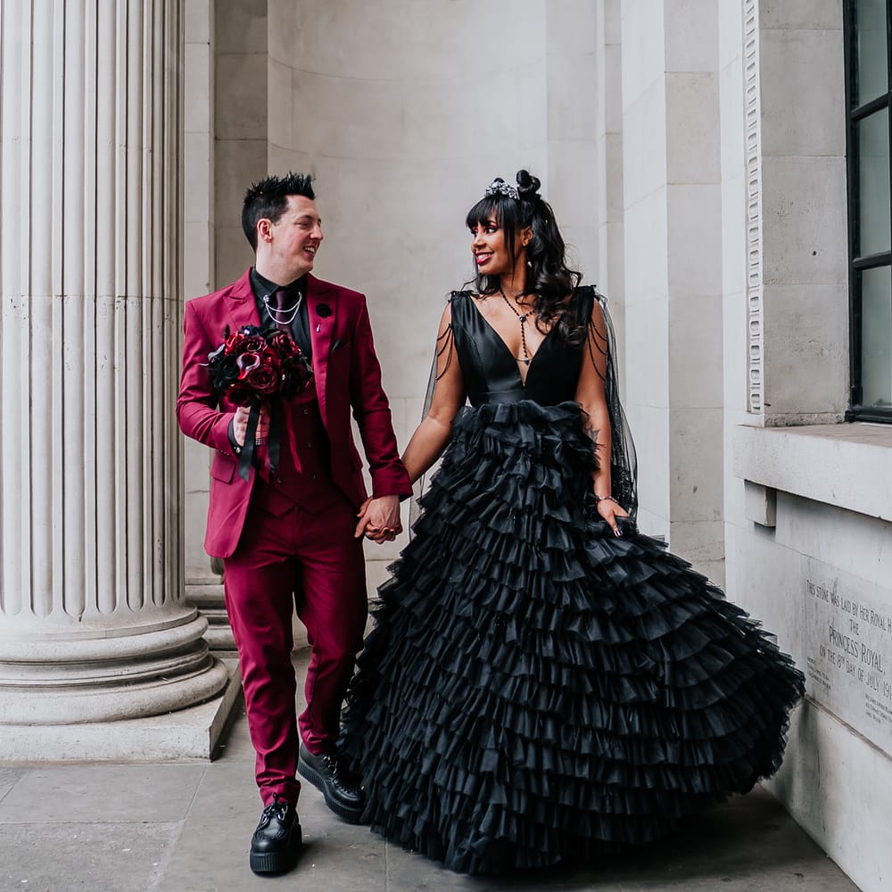 Bride wears black ruffled wedding dress and stands beside her groom wearing wine red suit with black shirt for couples portraits 