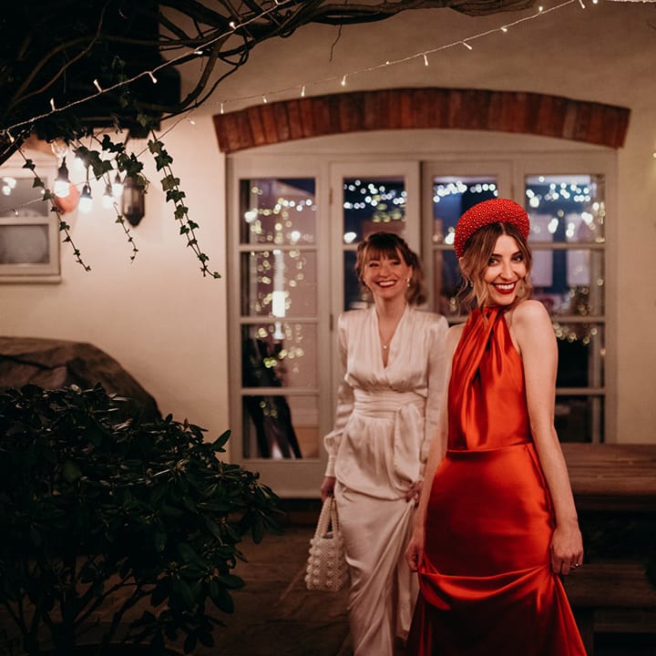 Bride in red silk wedding dress with cape and large red embellished head band standing with bridesmaid in ivory satin long sleeve bridesmaid dress