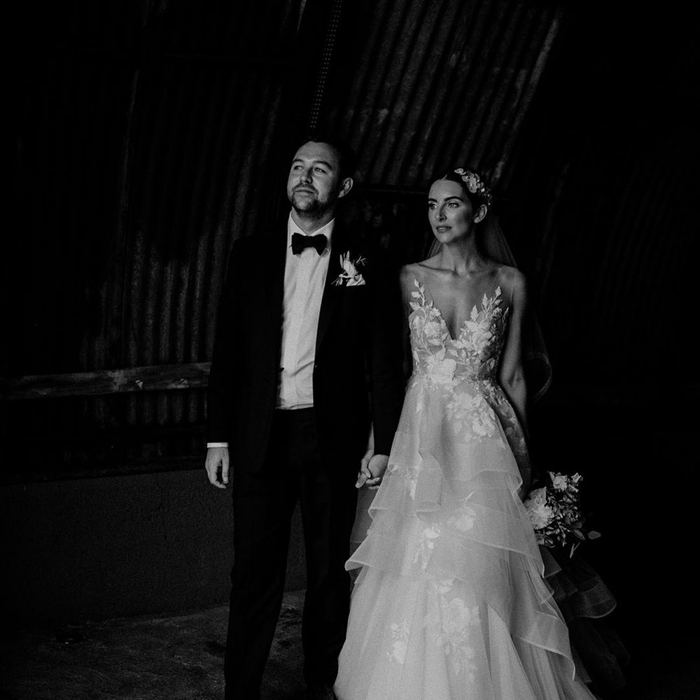 Groom in black tie stands holding hands with the bride in a flower layered tulle wedding dress and hairpiece 