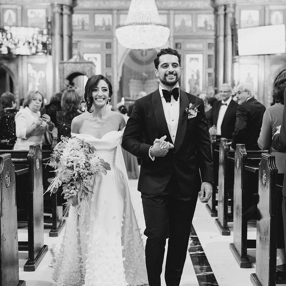 Bride in off the shoulder styled wedding dress walks down the aisle with her groom in black tie 