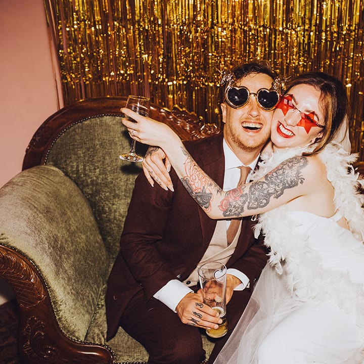 Bride & groom sit in DIY photo booth wearing fun sunglasses and gold streamers to the back