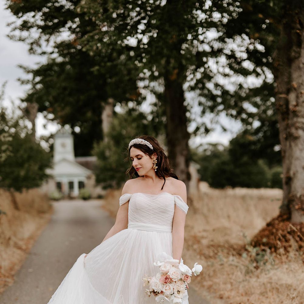 Bride in off the shoulder wedding dress wearing a pearl headband and statement earrings with neutral wedding bouquet 