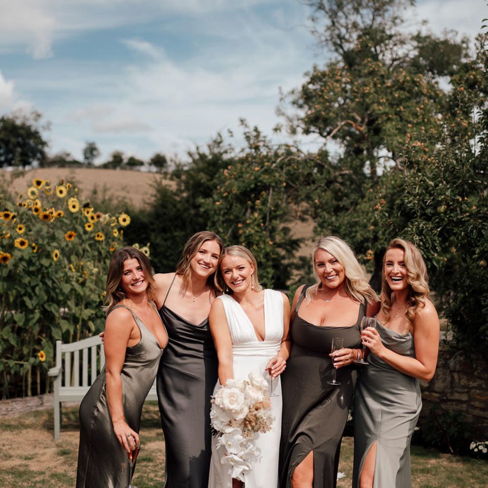Blonde bride with pearl earrings with plunging wedding dress holding a white orchid bouquet with the bridesmaids in mismatched dresses 