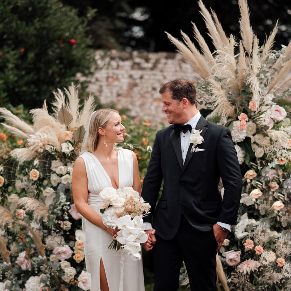 The bride and groom smile at each other standing in front of flower columns with pampas grass with pale pink and white roses 