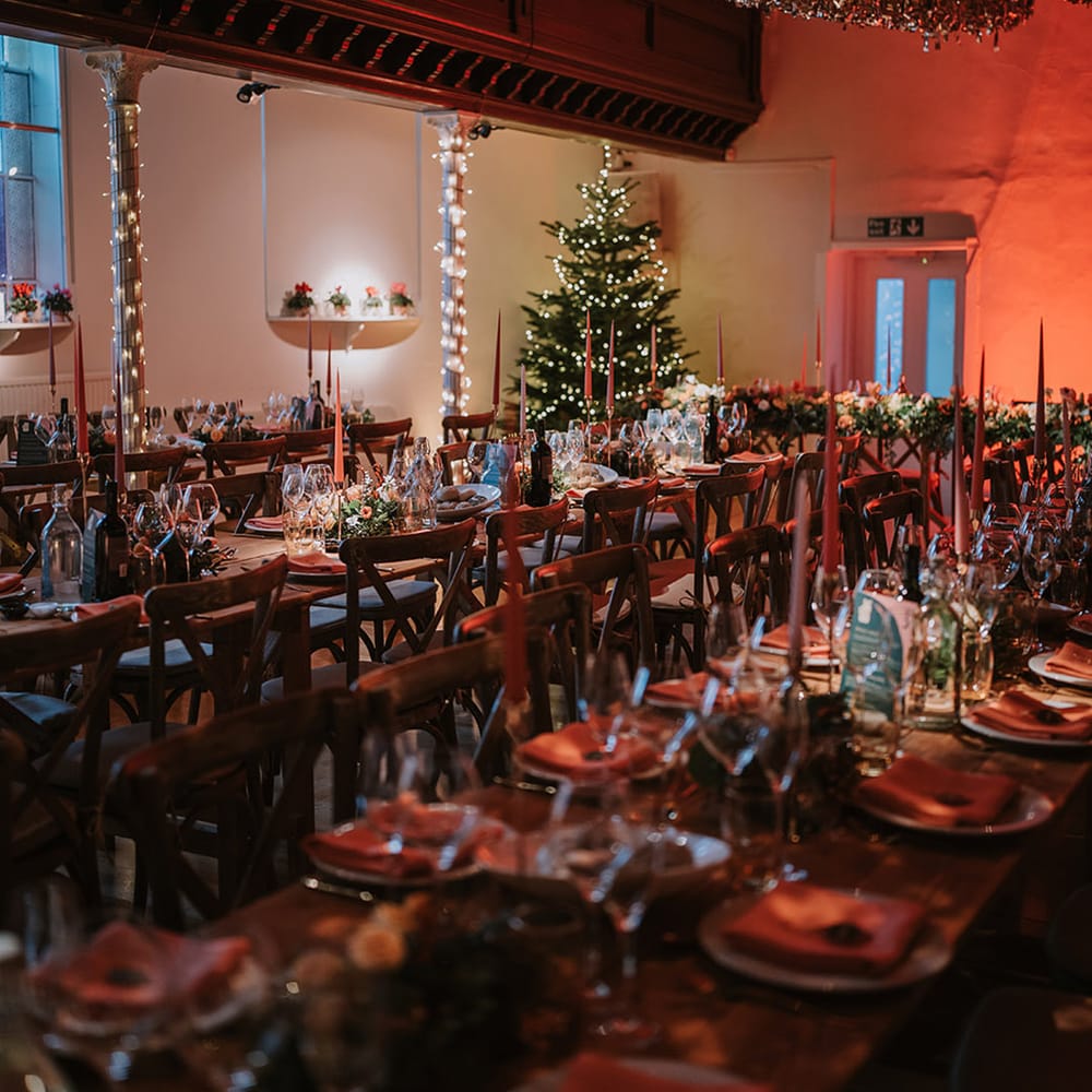 Reception room of Marylebone Town Hall with silver and gold wedding streamers, orange tapered candles, orange napkins, orange rose and foliage tablerunners, fairy lights and large Christmas tree 