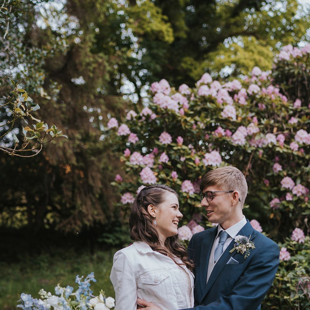 Bride in personalised white denim jacket smiling with the groom in navy suit at their woodland setting at Badgers Holt in Devon