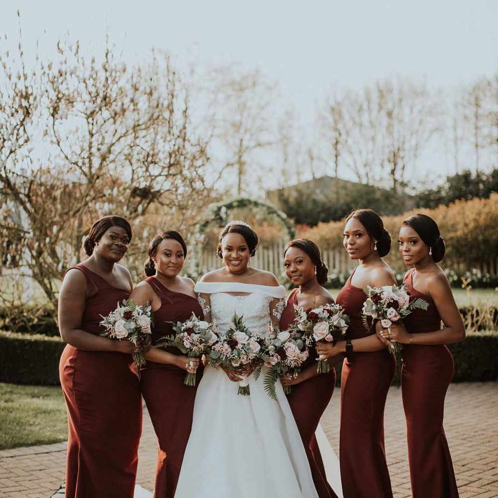 Bride in off the shoulder wedding dress posing with bridal party wearing one strap burgundy bridesmaid dresses 