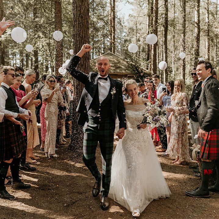 White paper wedding lanterns decorating the outdoor ceremony area with the bride in a floral wedding dress and groom in bespoke checkered wedding trousers