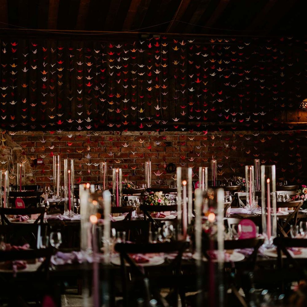 Origami paper cranes DIY wedding decor at Willow Marsh Farm Loughborough in front of rustic luxe wedding tablescape with long pink and purple tapered candles, tall candle glass jars, flower and foliage centrepieces and retro wedding signage 