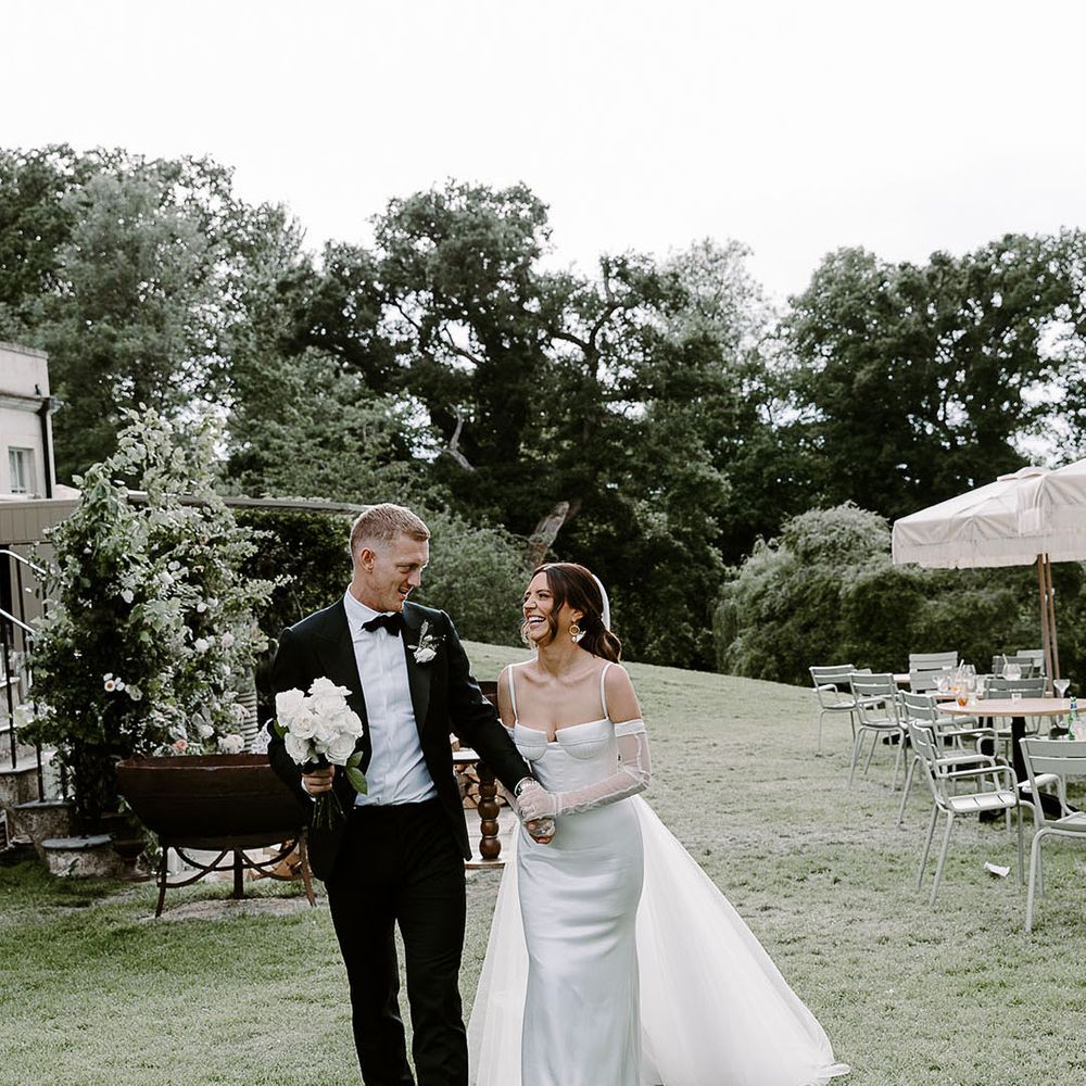 The groom in a black tuxedo walks with the bride in an Aleena Lena wedding dress 