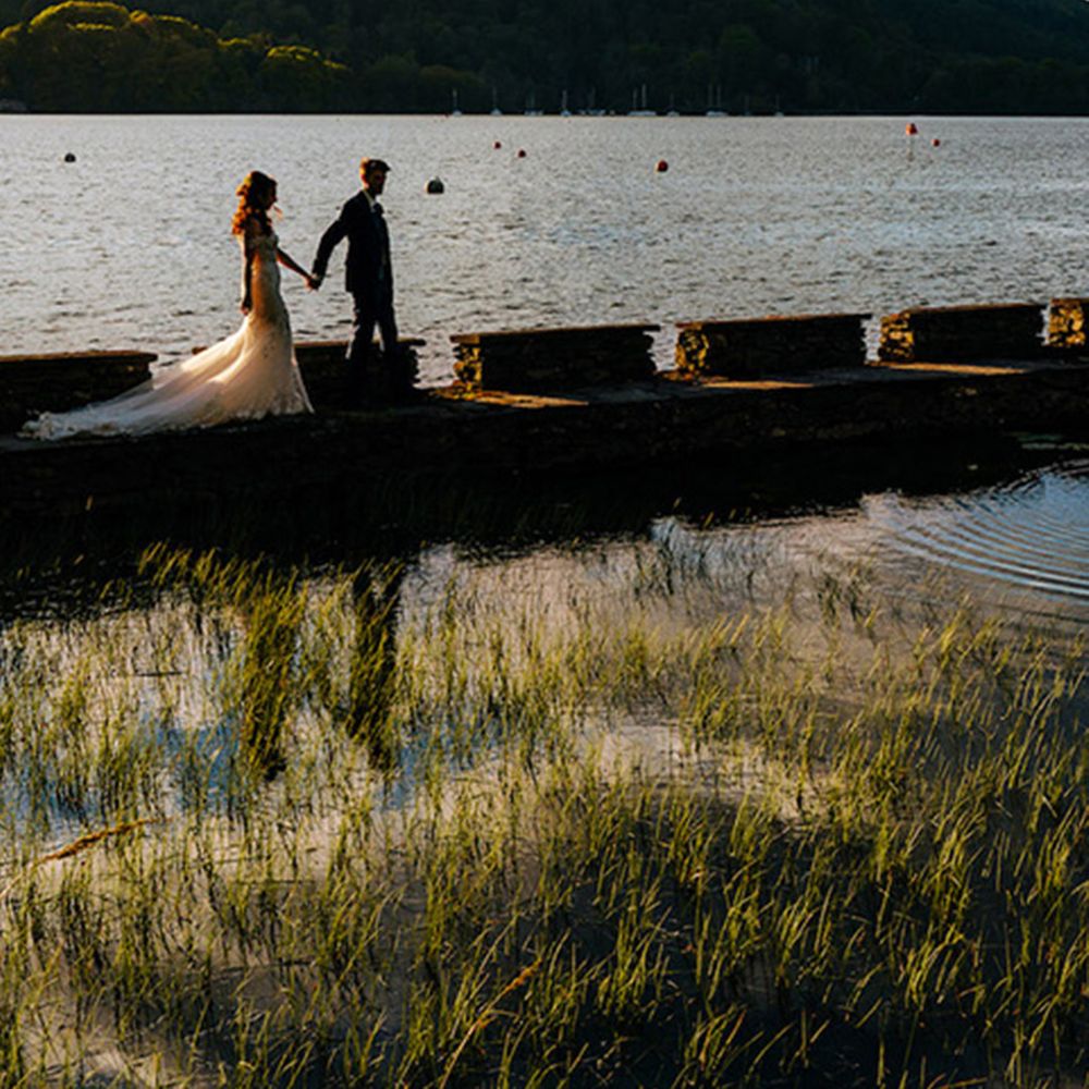 A bride in a mermaid wedding dress and groom walk across the jetty at the Lake District wedding venue 