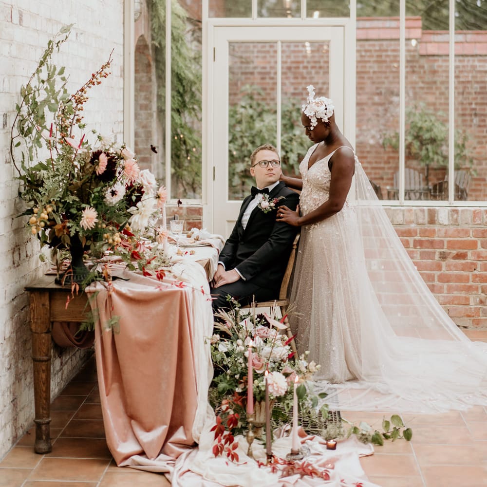 Bride with alopecia standing next to her groom at their intimate reception in a blush pink Julita Bridal London wedding dress with watteau train 