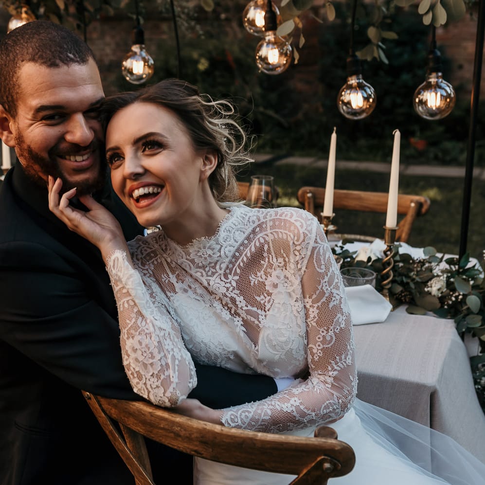 Black tie wedding with bride and groom laughing sitting at their festoon lit wedding reception table 