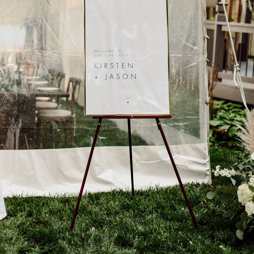 White wedding sign on easel reading 'Welcome to our love story Kirsten and Jason' outside white marquee at backyard wedding