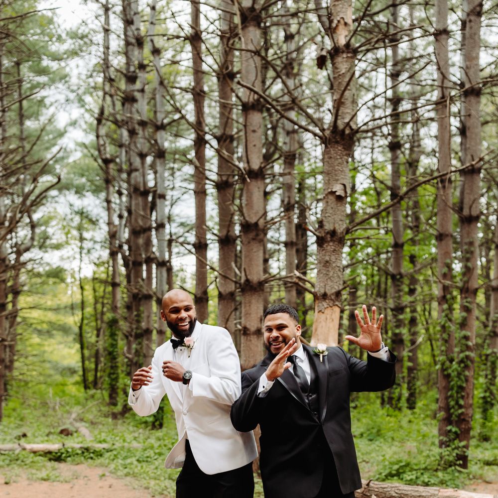 Groom in white tuxedo jacket jumps into air with groomsman in black suit in woodland at backyard wedding
