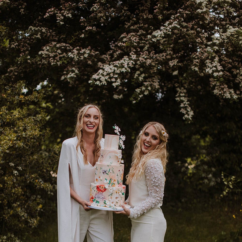 Two brides in a short wedding dress, bridal boots, jumpsuit and cape holding a wedding cake with flower illustration 