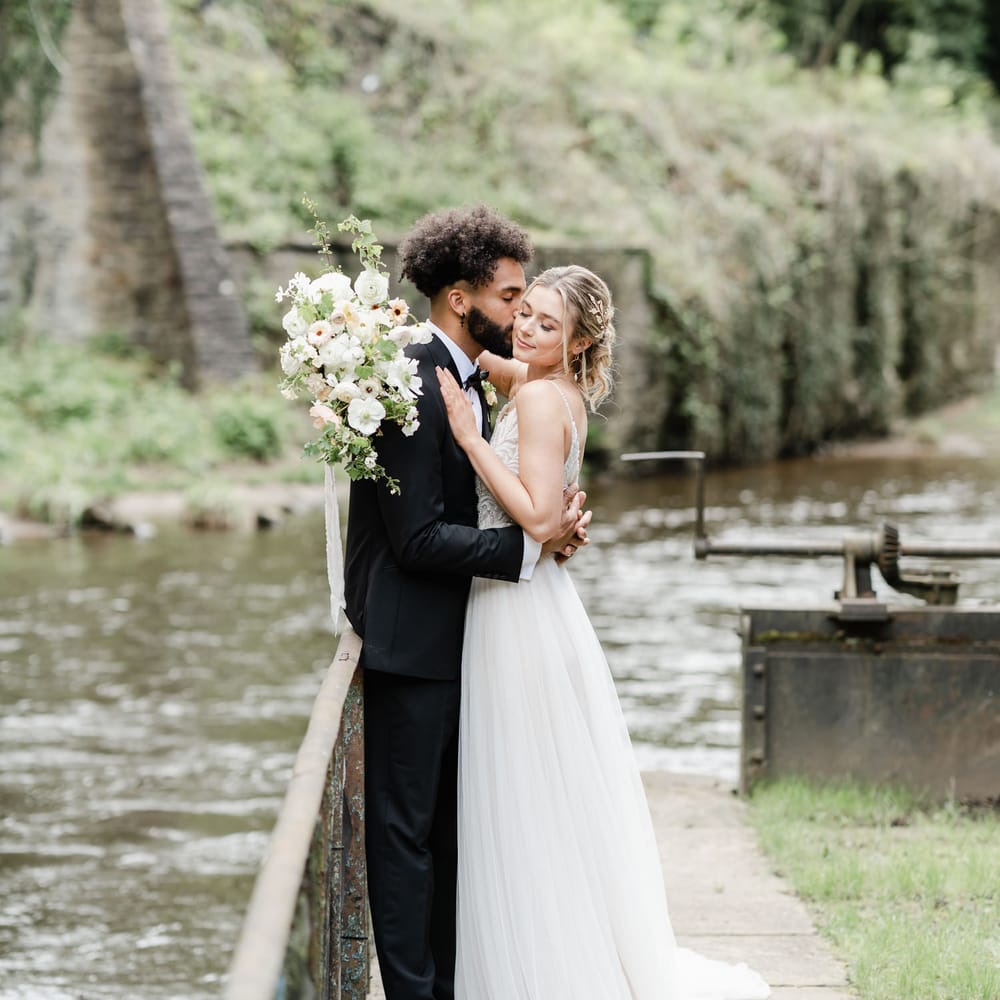 Groom in a tuxedo kissing his bride in a tulle skirt wedding dress by the banks of the River Goyt in New Mills Derbyshire