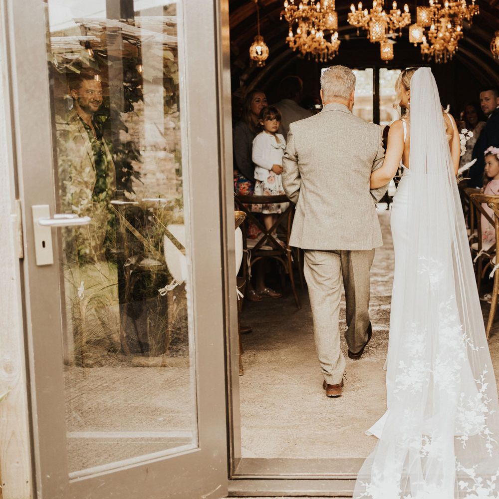 Bride in white Made With Love wedding dress and long embroidered veil walks into wedding ceremony room lit with chandeliers holding the arm of man in sage green suit at Anran Devon