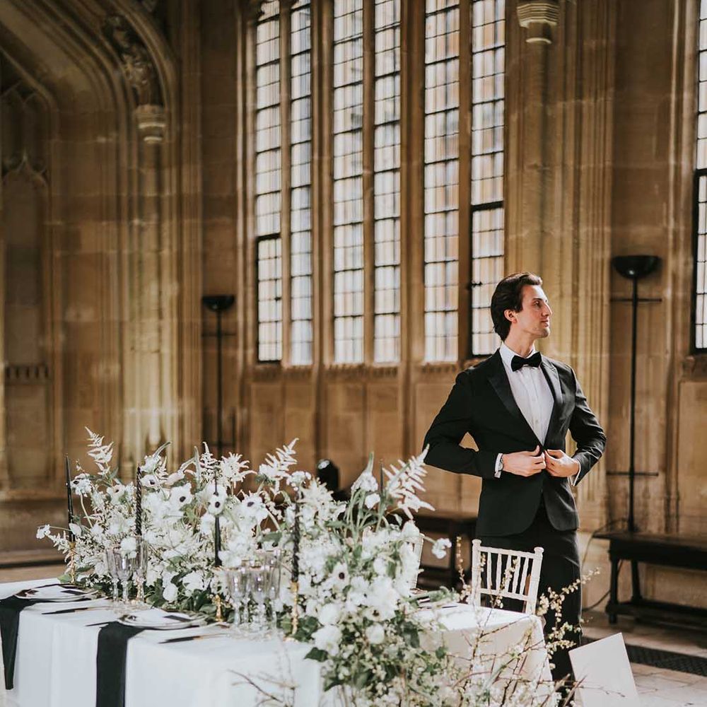 Groom in a tuxedo standing at the intimate reception table decorated with black, white and gold tableware and flowers 