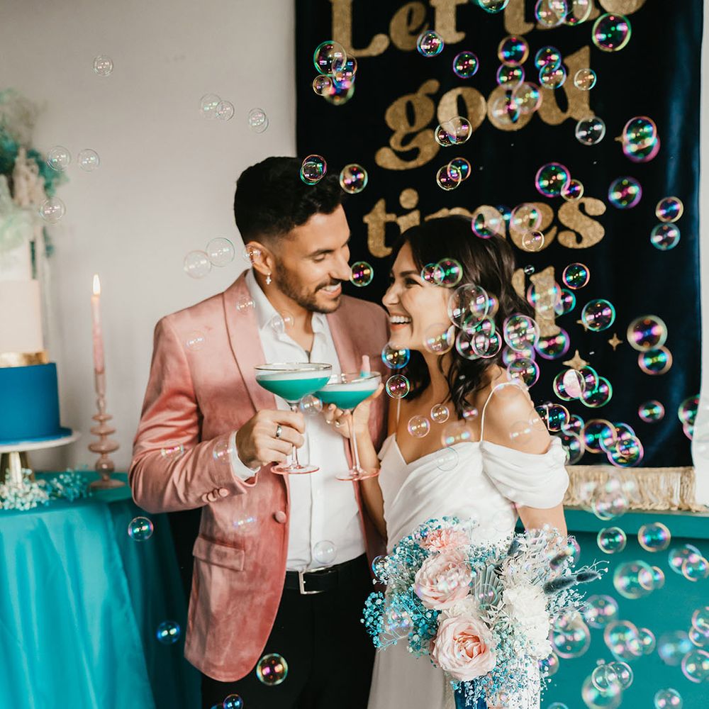 Groom in pink velvet jacket and bride in a cold shoulder wedding dress sipping cocktails with bubbles floating around them 