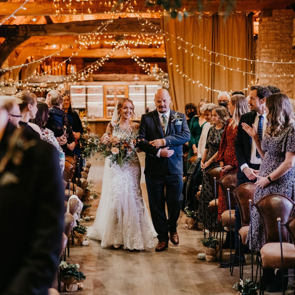 Bride in lace wedding dress with train and veil holding large mixed bridal bouquet walks down aisle with father during rustic barn wedding ceremony